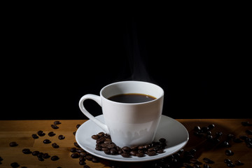 Brewed coffee in a white cup with steam or vapor and coffee beans scattered on a wooden surface and on the white saucer. 