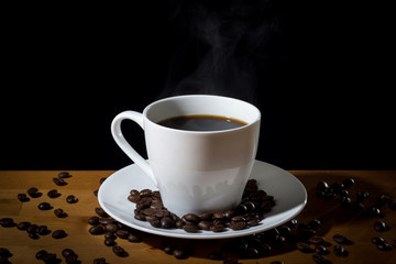 Brewed coffee in a white cup with steam or vapor and coffee beans scattered on a wooden surface and on the white saucer. 