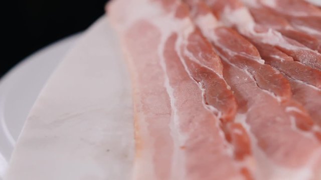 Layered fresh raw bacon sliced pieces on the rotating white cutting board. The close up shot of uncooked bacon strips on the clockwise rotating plate