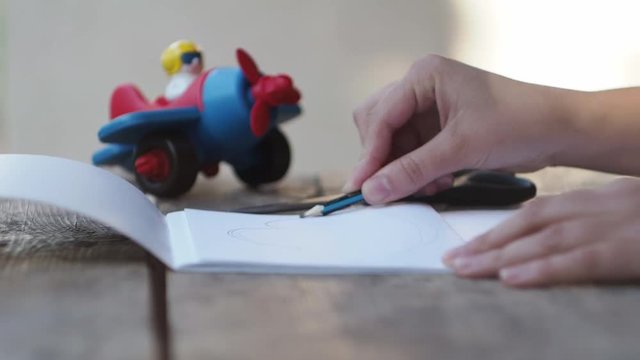 The woman draws a pencil on the sheet of paper. Close up. Wooden table. The toy plane with the pilot. Well lit background. Retro style. The artist behind work.