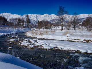 川岸より雪景色のアルプス、川には水が流れ、吊橋が架かり、古い民家、空は青く遠くには雪景色のアルプス