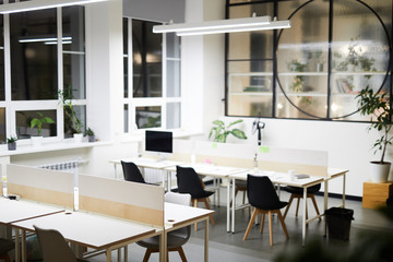 Modern open space office interior: black chairs at tables in rows under hanging lamp, potted plants near wall and on window sill
