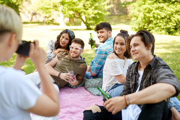 friendship, leisure and technology concept - woman with camera photographing his friends drinking non alcoholic drinks at picnic in summer park