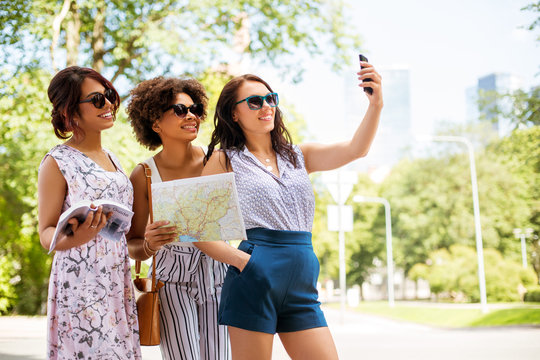 tourism, travel and technology concept - happy women with city guide and map taking selfie by smartphone in summer park
