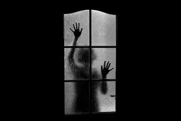 Dark silhouette of girl behind glass. Locked alone in room behind door on Halloween in grayscale. Nightmare of child with aliens, monsters and ghosts. Evil in home in monochrome. Inside haunted house.
