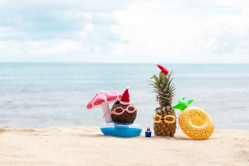  funny attractive pineapples and coconut in stylish sunglasses on the sand against turquoise sea. Wearing christmas hats. Christmas and new year vacation concept on tropical beach.