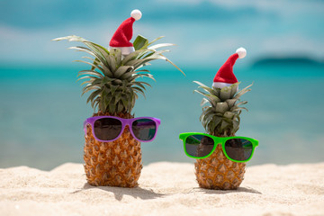 Family of funny attractive pineapples in stylish sunglasses on the sand against turquoise sea. Wearing christmas hats. Christmas and new year vacation concept on tropical beach. Family holiday. Bright