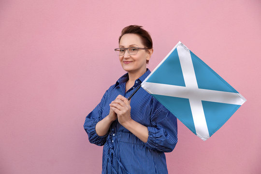 Scotland flag. Woman holding Scottish flag. Nice portrait of middle aged lady 40 50 years old holding a large flag over pink wall background on the street outdoors.