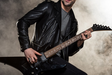 cropped view of rocker in leather jacket playing electric guitar on smoky background