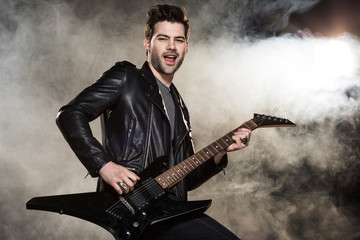 Fototapeta na wymiar handsome rocker in leather jacket playing electric guitar on smoky background