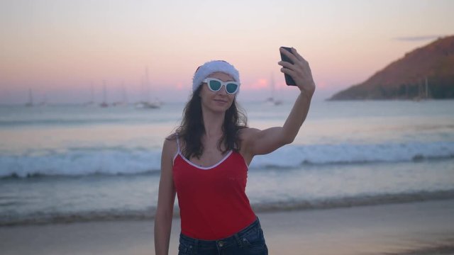 Fun woman in red santa hat taking selfie photo at beach vacation. Summer holiday girl happy at smartphone camera taking self-portrait on her travel vacations on pristine paradise beach.