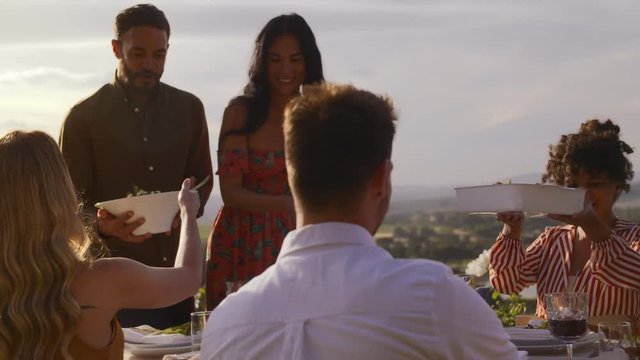 Couple serving food to friends at party. Group of men and women having dinner together outdoors.