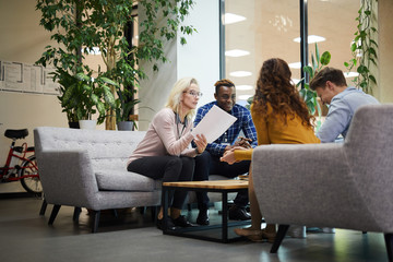 Confident mature lady in glasses sitting on sofa in lobby and reading report to colleagues while sharing ideas for new project at meeting