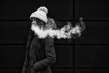 Vape teenager. Young pretty white girl smoking an electronic cigarette opposite modern background on the street in the winter. Bad habit. Black and white.