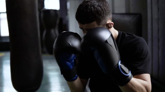 Handhelded footage of young boxer's training. Dark haired man hitting the boxing bag, hard kicks. Motivation in sport. Old style gym, daytime. Close up