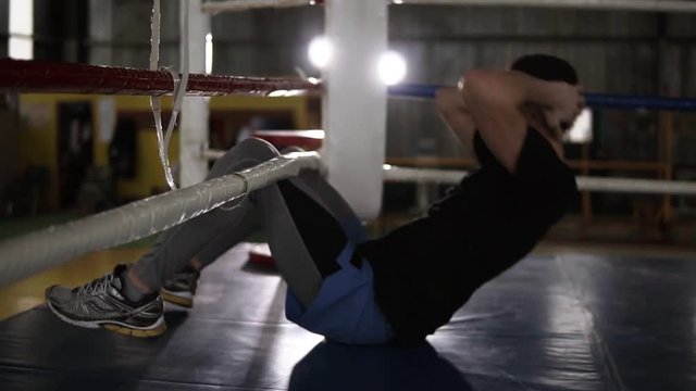 Boxer sitting on the floor at the boxing ring training abdominal muscles. Lifting torso up to the boxing ropes. Training process at boxing gym. Side view