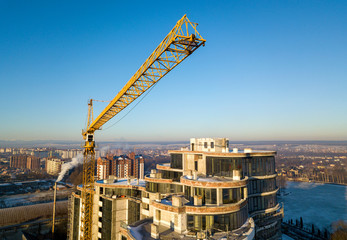 Apartment or office tall building under construction, top view. Tower crane on bright blue sky copy space background, city landscape stretching to horizon. Drone aerial photography.