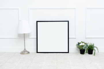 There are beautiful flowers and various objects near the rectangle blank picture frame in front of white wall on the carpet living room.