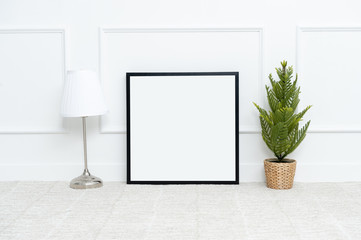 There are beautiful flowers and various objects near the rectangle blank picture frame in front of white wall on the carpet living room.