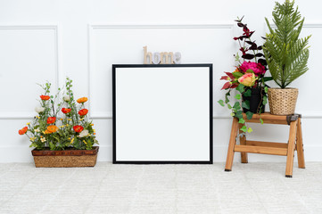 There are beautiful flowers and various objects near the rectangle blank picture frame in front of white wall on the carpet living room.