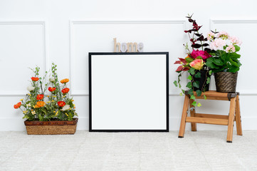 There are beautiful flowers and various objects near the rectangle blank picture frame in front of white wall on the carpet living room.