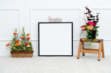 There are beautiful flowers and various objects near the rectangle blank picture frame in front of white wall on the carpet living room.