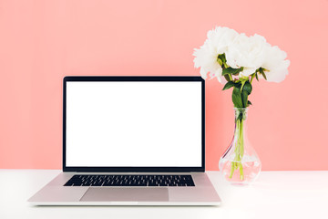 Laptop with white blank screen and flowers in a vase on a table on a coral background. mock up