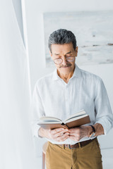 senior man in glasses standing and reading book at home