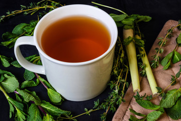 Herbal tea in a white mug on a dark background. Fresh herbs around: mint, thyme and lemon sargo.