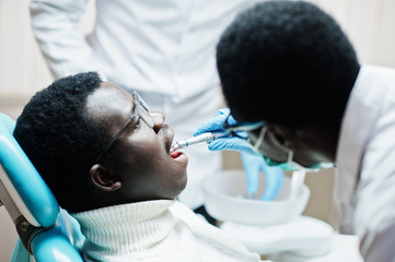 Fototapeta premium African american man patient in dental chair. Dentist office and doctor practice concept. Professional dentist helping his patient at dentistry medical. Drilling patient's teeth in clinic.