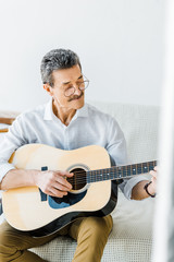 cheerful senior man in glasses playing acoustic guitar at home