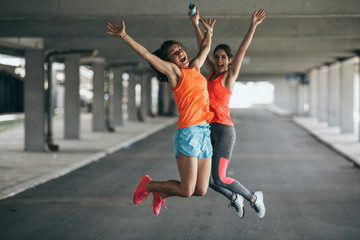 Two young female runners jumps on the street, expressing positive emotion. Fitness and workout concept.	