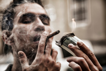 Young brutal businessman in the smoke lighting cigar with $100 dollar bill. Selective focus on...