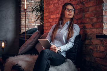 Beautiful young girl dressed in a stylish shirt holds the tablet sitting on the couch and looks out the window in the room with a loft interior.