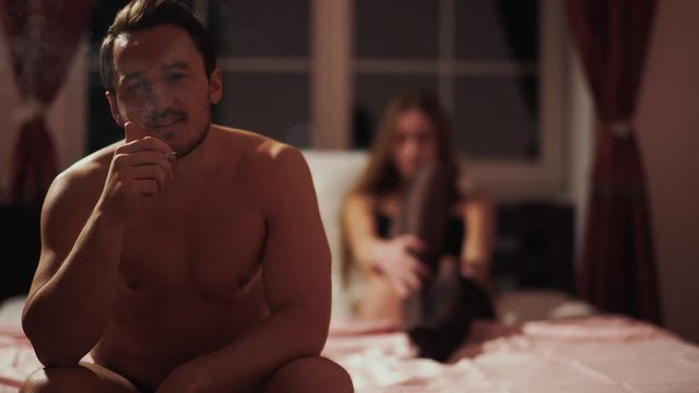 Happy guy smokes sitting on the bed on the backgrund of a beautiful young girl in stockings