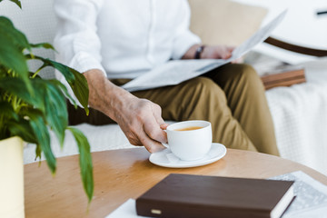 cropped view of retired man holding cup with coffee and newspaper at home