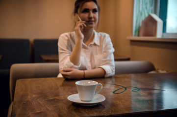 girl sits in a cafe, looks out the window, holds the phone, waits, a white cup of coffee in the foreground on the table