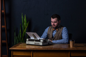 Bearded stylish writer typing on typewriter. Modern writer working on new book in office. Dark background. Space for text