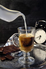 Milk from the milk jar is poured into a cappuccino latte coffee and beautifully mixed with coffee in a glass cup beside a chocolate bar. Dark atmospheric food photo.milk jar,dark food foto,latte coffe