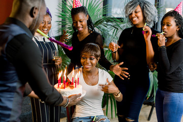 Portrait of charming African American girl blowing on candles on birthday cake surrounded by friends at party