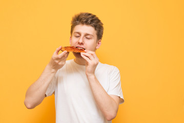 Young man eats a piece of appetizing pizza on a yellow background. Hungry guy holds a piece of pizza in his hands, looks at him and is going to eat it, isolated on a yellow