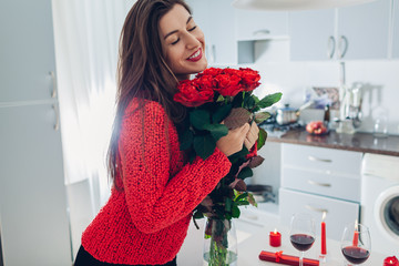 Young woman found red roses with candles, wine and gift box on kitchen. Happy girl holding flowers. Valentines day