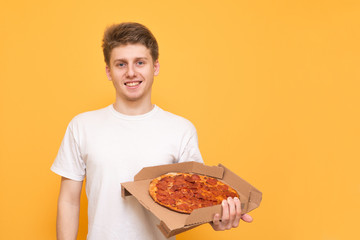 Positive guy in a white T-shirt,isolated on a yellow background,holds a box of pizza in his hands, looks into the camera and smiles. Fast Food Concept.