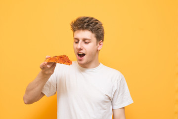 Portrait of a hungry guy with a piece of pizza in his hands, isolated on a yellow background. Young man stands on a yellow background and is focused on a piece of pizza in his hands.