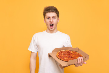 Amazed young man in a white T-shirt is isolated on a yellow background, holds a box of pizza in his hands, looks into the camera. Pizza Concept. Discount on a pizza