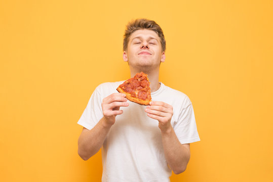 Portrait Of A Guy In A White T-shirt Eats A Piece Of Fresh Pizza And Gets A Pleasure, Isolated On A Yellow Background. Young Man Is Happy To Eat A Delicious Piece Of Pizza On A Yellow Background.