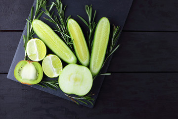 Green fruits, vegetables and rosemary on black background with copy space. Creative figure of natural green products. Cucumbers, kiwi, lime and green apple on slate board