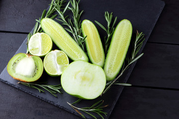 Green fruits, vegetables and rosemary on black background with copy space. Creative figure of natural green products. Cucumbers, kiwi, lime and green apple on slate board