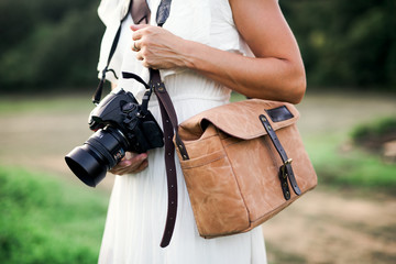 A midsection of woman in nature holding a camera and a brown leather bag.