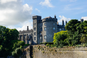 Fototapeta na wymiar Fortified castle with grey stone walls surrounded by green trees under a blue sky with white clouds. Kilkenny Castle in Ireland.